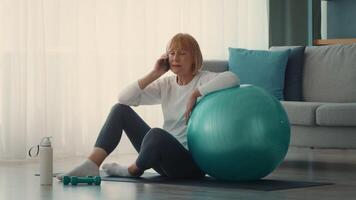 An elderly woman engages in a workout routine at home, seated on a yoga mat next to an exercise ball, talking on her phone, showcasing an active lifestyle. video