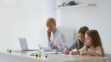 A mother on a phone call with two daughters drawing and coloring at a table. They are enjoying their family time in a bright, cozy home setting during the day. video