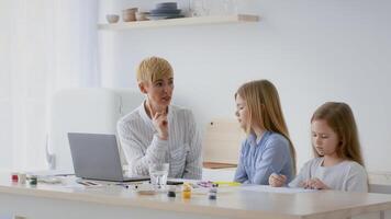 A teacher engages with two children at a kitchen table, discussing activities while the kids draw and write. Natural light fills the room, enhancing the warm, inviting atmosphere. video