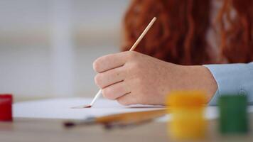 A teenager with red hair is focused on painting at home. She is seated in a wheelchair, using a brush to create art on paper. The atmosphere is calm and creative. video