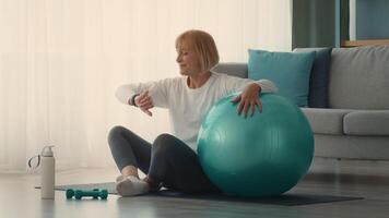 A retired Caucasian woman is exercising at home using a large blue exercise ball. She appears focused as she incorporates fitness into her daily routine in a modern living space. video