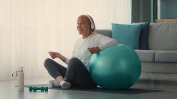 A cheerful elderly lady engages in a workout session at home, using a stability ball for exercise. She wears headphones and enjoys her fitness routine in a bright, cozy space. video