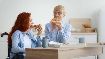 A redhead girl in a wheelchair smiles while sharing pizza with a friend in a bright, modern kitchen. They are engaged in a cheerful conversation, holding slices of pizza. video