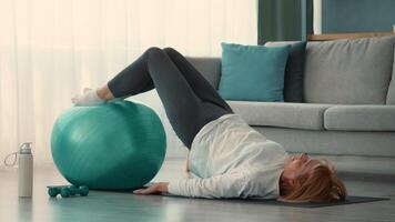 A senior woman is exercising at home, balancing on an exercise ball while working on her fitness routine. She maintains a focused expression, promoting healthy living. video