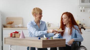 A teenager in a wheelchair enjoys a joyful occasion at home as someone serves her a small cake. Decorations and gifts are placed on the table, creating a festive atmosphere. video