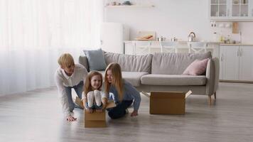 A family is having fun together in a bright living room, where a child is sitting inside a cardboard box while mother and sister help push the box. The space is cozy and welcoming. video