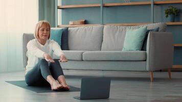 A senior woman engages in a workout session at home, following exercises on a laptop. She sits on a mat, focused, in a living room that features a cozy sofa and simple decor. video