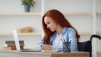 A redhead girl in a wheelchair sits at a desk, focused on her laptop while taking notes. Shelves in the background hold books, creating a cozy home study environment. video