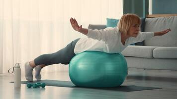 An elderly woman practices a workout routine on a stability ball in her cozy living room. She focuses on her form while promoting fitness and well-being at home. video