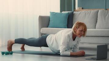 A retired woman engages in a workout routine in her home, focusing on strength and balance. She exercises on a mat, following a digital guide, showcasing active senior life. video