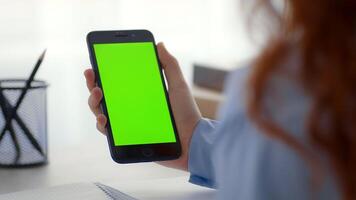 A teenage girl sits at home, holding a smartphone displaying a bright green screen. She has long red hair and appears focused while interacting with the device. The environment is cozy and well-lit. video