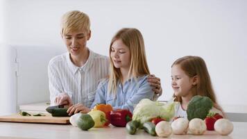 A woman is guiding two young girls as they prepare fresh vegetables for a healthy meal. The kitchen is bright, with a variety of colorful produce on the table. video