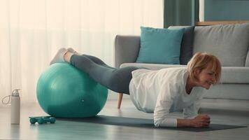 An elderly woman performs an exercise routine using a stability ball in her living room. She focuses on maintaining her balance and strength, showcasing a dedication to fitness in her retirement. video