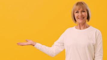A smiling woman in a white top extends her arm with an open hand, showcasing an inviting gesture. The vibrant yellow backdrop enhances her warm demeanor and cheerful expression, copy space video