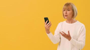 A woman with short hair is animatedly speaking into her smartphone, displaying emotions while standing in front of a bright yellow background. She appears engaged and expressive during the call. video