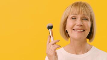 A smiling older woman uses a makeup brush to apply blush to her cheeks. She stands in front of a vibrant yellow background, highlighting her joyful expression and beauty routine, copy space video