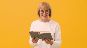 A cheerful older woman with glasses smiles while holding an open book in front of a vibrant yellow background, showcasing her enjoyment of reading and learning. video
