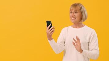An older woman smiles and waves while enjoying a call on her smartphone, set against a vibrant yellow background, showcasing joy and connection in her communication, copy space video
