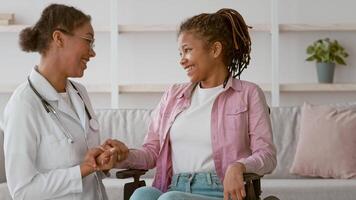 A healthcare professional smiles while holding the hand of a patient in a wheelchair. The warm environment suggests a supportive atmosphere for recovery and conversation. video