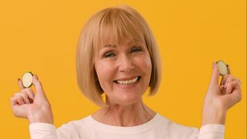 An elderly woman smiles brightly while playfully holding cucumber slices in front of her eyes. The vibrant yellow background adds a cheerful atmosphere to the lighthearted moment. video