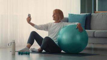 A senior woman engages in her fitness routine at home, taking a moment to enjoy her workout. She uses an exercise ball and captures the moment on her phone, showcasing an active lifestyle. video
