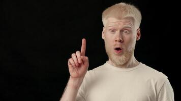 A young man with white skin and hair stands against a dark background, displaying a thoughtful expression. His appearance highlights the distinct characteristics of albinism, copy space video