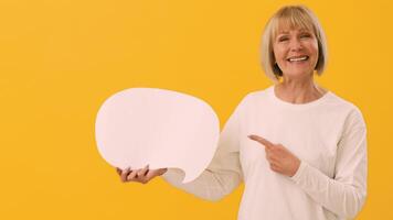 A smiling woman in a white long-sleeve shirt points to a blank speech bubble in a vibrant yellow studio, suggesting a conversation or message creation. The atmosphere is lively and inviting. video