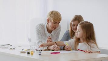 A caregiver engages with two young girls as they joyfully create art at a table. The bright, airy room provides an inspiring atmosphere for their colorful drawings. video