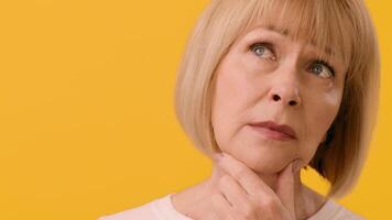 A senior woman with a bob haircut appears deep in thought, resting her chin on her hand while facing a vibrant yellow backdrop. Her expression suggests introspection and contemplation, copy space video