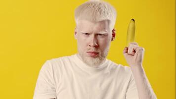 A young man with albinism aims a banana playfully at the viewer. His white hair and skin contrast sharply with the vibrant yellow backdrop, creating a striking visual. video