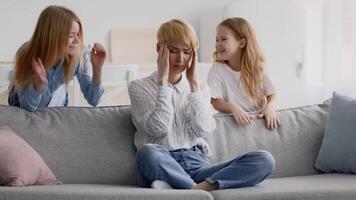 A young adult appears overwhelmed while seated on a sofa, as two children stand playfully beside him. The setting is a cozy living room filled with natural light. video