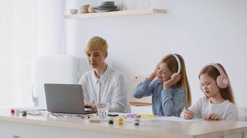 A mother assists her two children as they engage in creative activities at a bright kitchen table. One child listens to music while the other draws, creating a warm, productive atmosphere. video