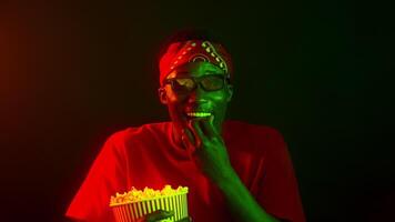 A stylish African American man smiles joyfully while holding a bucket of popcorn in a vibrant neon-lit environment. His energetic expression adds to the cool atmosphere. video
