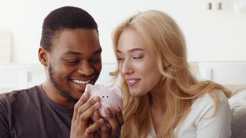 A happy biracial couple is sitting together in their cozy home, showcasing their love and affection. They are playfully holding a pink piggy bank, symbolizing their future together. video