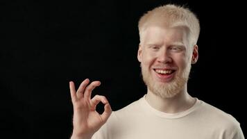 A young man with albinism features striking white hair and skin. He stands indoors against a dark background, displaying a thoughtful expression and unique style, copy space video