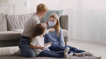 A caregiver gently assists a child in adjusting her face mask while another child sits nearby, all in a cozy living room filled with natural light. video