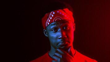 A cool millennial man poses against a dark backdrop illuminated by neon lights. He wears a vibrant bandana and displays a thoughtful expression, embodying a modern, hipster aesthetic. video