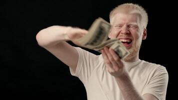 A young man with albinism is joyfully sorting through a stack of cash while smiling. He has white skin and hair, making him stand out in the studio environment. video