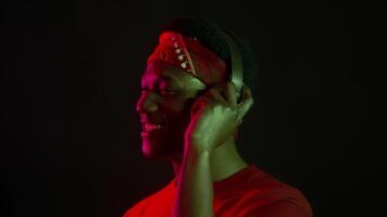 A handsome African American man wearing a red bandana and a matching shirt smiles while listening to music with headphones in a neon-lit, isolated environment, creating a futuristic vibe. video