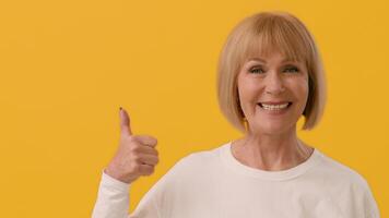 A cheerful woman with a short blonde bob hairstyle wears a white shirt while standing in front of a vibrant yellow background, exuding warmth and positivity in her expression, copy space video