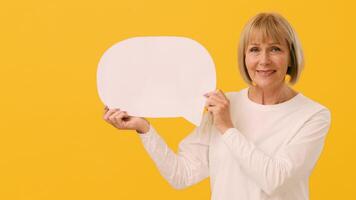 A woman with shoulder-length blonde hair is smiling gently while holding a blank speech bubble. She stands in front of a vibrant yellow backdrop, inviting conversation or expression. video