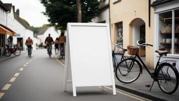 Peaceful small-town street with a white empty sandwich board. A craft store and bicycles set the scene, while blurred figures in the background add life. photo