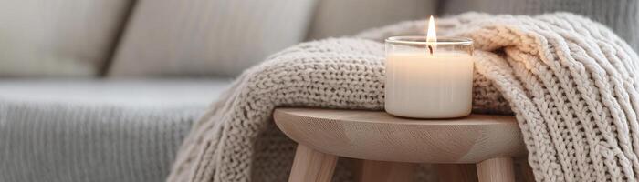 A lit candle sits gracefully on a small wooden table, accompanied by a folded knitted blanket in muted gray. This cozy setting exudes warmth against a soft neutral backdrop photo