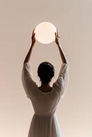 A confident woman stands with her back to the viewer, lifting a glowing globe above her head. Subtle rays of light surround her against a neutral gradient backdrop, creating an inspiring atmosphere photo