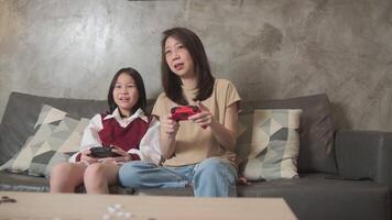 Asian family, mum, and kid are playing VDO game with joy controllers on sofa in home living room together, enjoy and cheerful, happy weekend exciting hobby of child and parent, digital entertainment. video