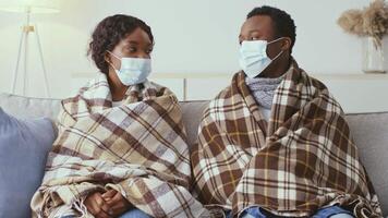 A millennial couple sits close together on a sofa, wrapped in warm blankets while wearing masks. They appear to be unwell, likely suffering from flu-like symptoms at home. video