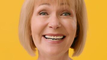 An older woman with a short blonde haircut smiles warmly, showcasing her bright teeth against a cheerful yellow backdrop. Her joyful expression conveys positivity and warmth. video