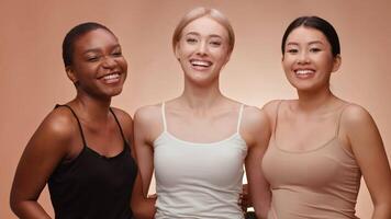 Three women of different backgrounds are smiling and posing closely together. They wear simple, neutral-colored tops and showcase a joyful atmosphere in a well-lit indoor space. video