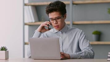man fokuserade på hans arbete i en avlägsen miljö engagerar i en telefon konversation medan skriver på hans bärbar dator. hyllor i de bakgrund innehålla böcker och växter, skapande en produktiv miljö. video