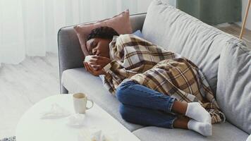 A young African American woman rests on couch, wrapped in a blanket while dealing with illness. Surrounded by tissues, illustrating the struggle with flu symptoms during isolation. video